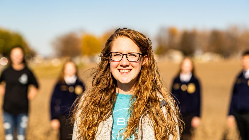 An student with wavy hair wearing glasses and smiling in the foreground, 4 kids wearing navy blue FFA jackets standing ing the background